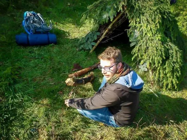 Mann sitzt vor einer improvisierten Tarp-Notunterkunft und bearbeitet Holzstücke
