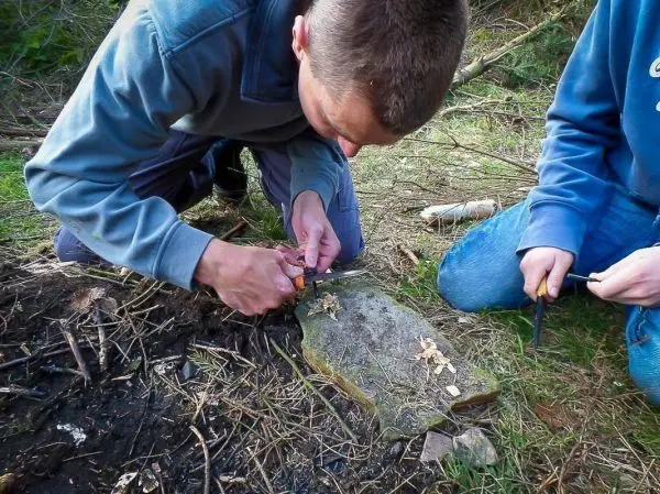 Messer schnitzt Zunder auf einem Holzstück neben einem kleinen Bodenfeuer