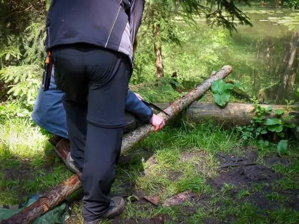 Person schneidet einen Baumstamm am Ufer eines Gewässers