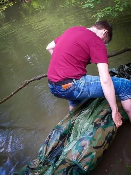 Person sitzt auf einer camouflagefarbenen Plane im Wasser und balanciert auf einem Ast