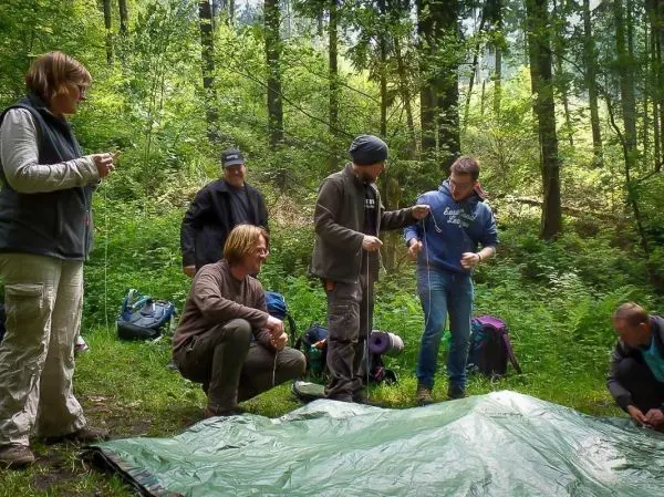 Sechs Personen arbeiten gemeinsam an der Aufstellung eines Tarps im Wald