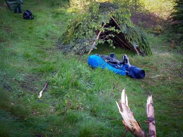 Tarp-Notunterkunft aus Ästen und Blättern neben einem blauen Rucksack auf Gras