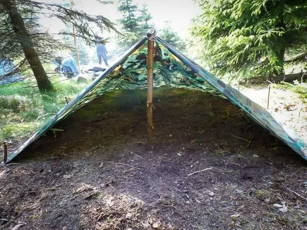 Tarp-Notunterkunft mit Holzstütze und Heringen im Wald aufgebaut