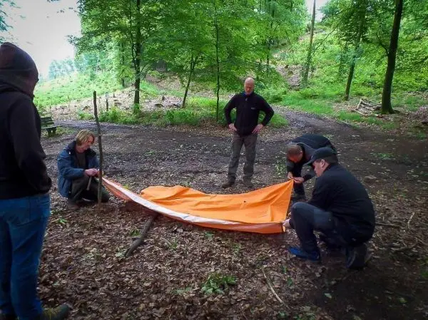Tarp-Notunterkunft wird von mehreren Personen aufgebaut und mit Stöcken gesichert