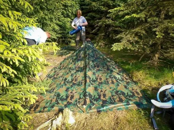 Tarp-Notunterkunft wird zwischen Bäumen aufgestellt, während eine Person Ausrüstung vorbereitet