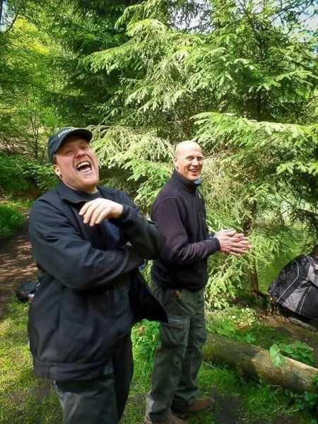 Zwei Männer lachen und stehen in einem Waldgebiet neben einem Gewässer