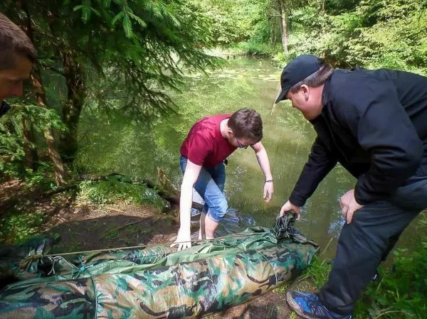 Zwei Personen heben eine camouflagefarbene Tarp-Notunterkunft am Ufer eines Gewässers