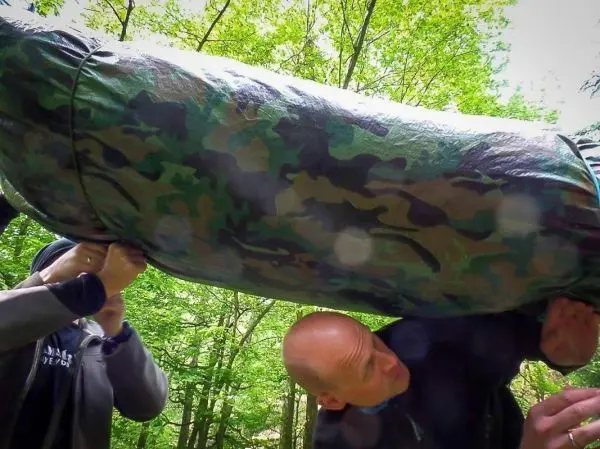 Zwei Personen heben eine camouflagefarbene Tarp-Notunterkunft über ihren Köpfen