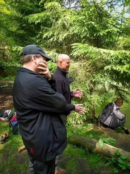 Zwei Personen stehen am Ufer eines Gewässers, eine Person sitzt am Boden