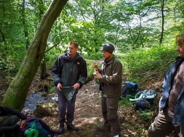Zwei Personen stehen im Wald und sprechen miteinander, Rucksäcke liegen daneben