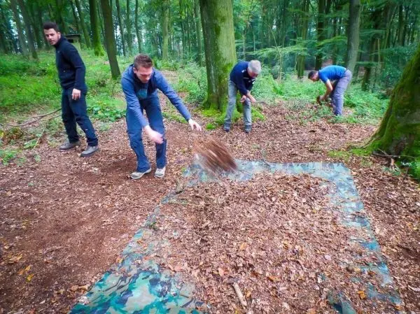 Blätter werden auf eine camouflagefarbene Plane geschaufelt, während mehrere Personen arbeiten