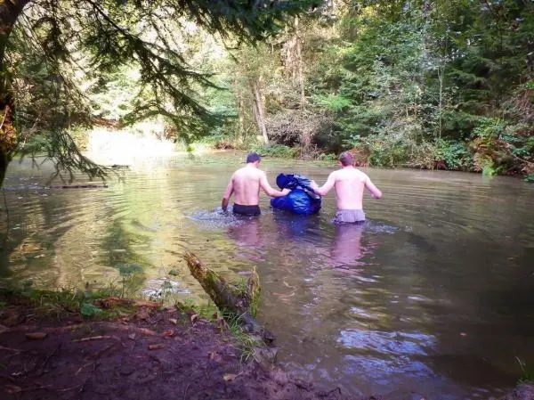 Drei Personen waten mit einem blauen Sack durch einen Fluss im Wald