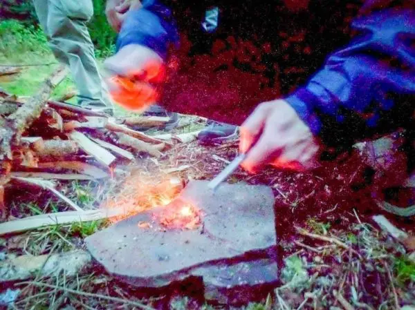 Feuerstahl erzeugt Funken auf Zunder, während Holzstapel im Hintergrund liegen