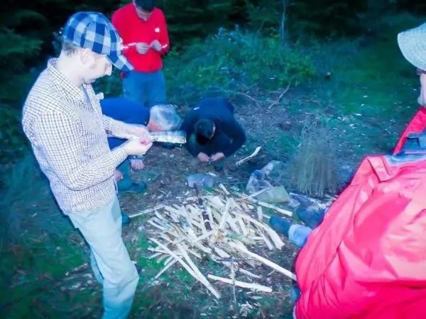 Mann mit Messer bereitet Zunder aus Holzstücken vor, andere Personen arbeiten am Boden