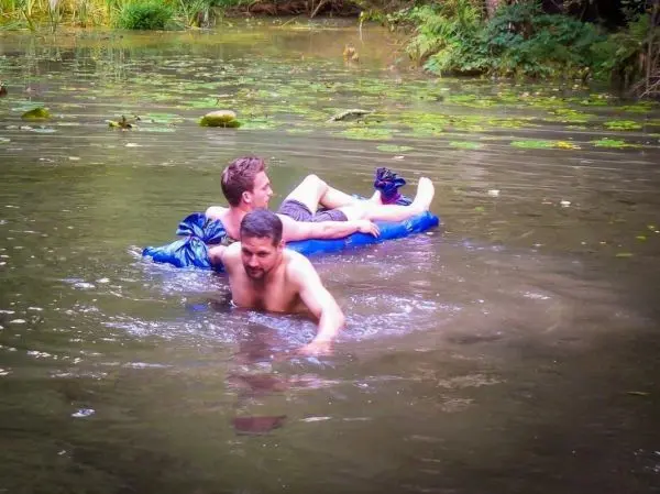 Zwei Personen im Wasser, einer schwimmt, der andere liegt auf einer blauen Luftmatratze
