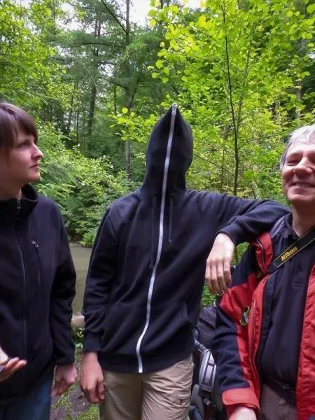 Drei Personen stehen im Wald, eine Person trägt einen Kapuzenpullover mit geschlossenem Reißverschluss