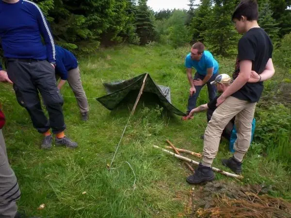 Gruppe baut Tarp-Notunterkunft mit Stöcken und Abspannschnur im Wald