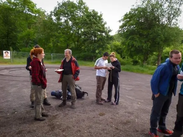 Gruppe steht auf einem Schotterplatz, einige halten Essen und schauen auf Zettel