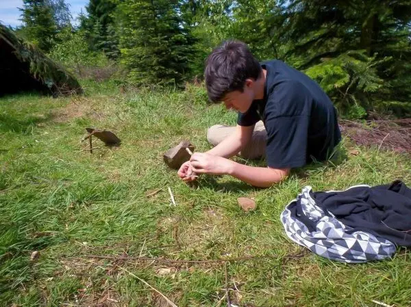 Junge baut eine Schlagfalle (Figure-Four) aus Holzstücken und Steinen