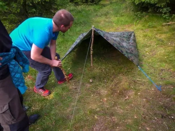 Mann befestigt eine Tarp-Notunterkunft mit einem Stock und Abspannschnur