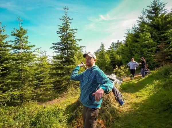 Mann mit Kapuze zeigt auf einen Weg zwischen Nadelbäumen, zwei Personen im Hintergrund