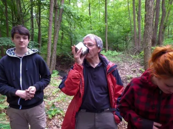Person trinkt aus einer Tasse im Wald, zwei weitere Personen stehen daneben