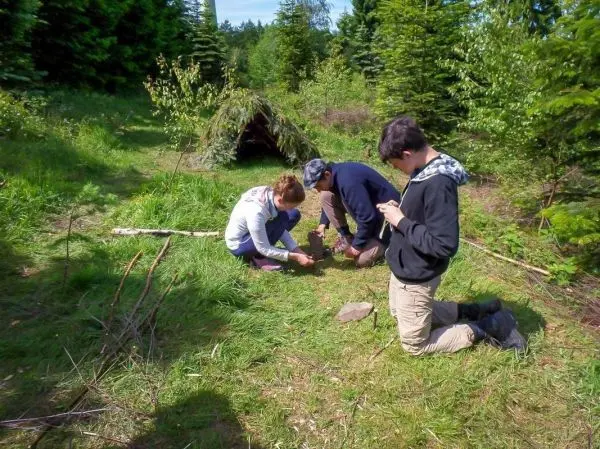 Zwei Personen bereiten Zunder auf dem Boden vor, während eine dritte Person beobachtet