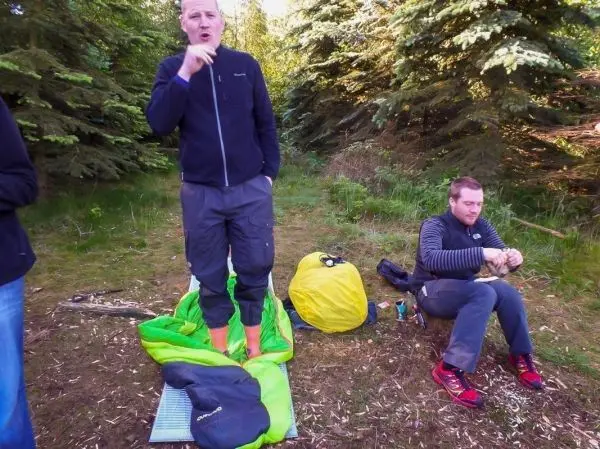 Zwei Personen in einem Wald, eine steht auf einer Isomatte, die andere sitzt und isst