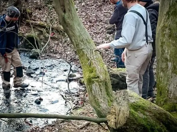 Drei Personen stehen am Ufer eines kleinen Gewässers und betrachten den Boden