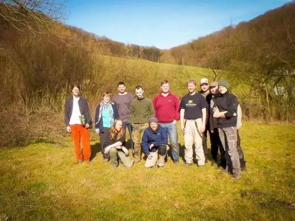 Gruppe von zehn Personen steht auf einer Wiese vor bewaldetem Hang