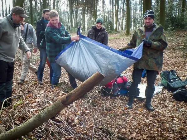 Gruppierung baut Tarp-Notunterkunft mit einer Plane und Holzstücken im Wald auf