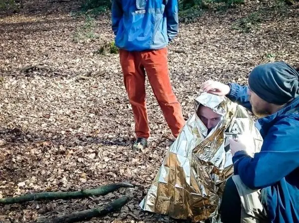 Mann in Rettungsdecke erhält Erste Hilfe im Wald, während eine andere Person beobachtet