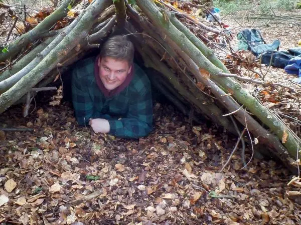 Mann kriecht aus einer selbstgebauten Laubhütte auf dem Boden