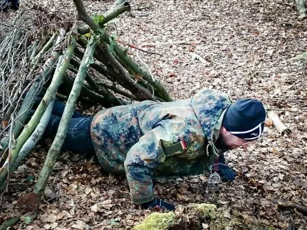 Mann kriecht unter einer selbstgebauten Laubhütte im Wald