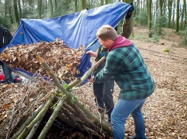 Person deckt eine selbstgebaute Unterkunft mit Laub ab