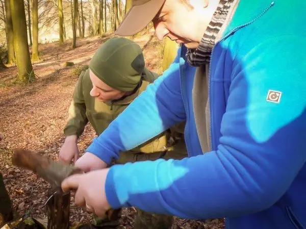 Zwei Personen bearbeiten einen Holzstock mit einem Messer im Wald