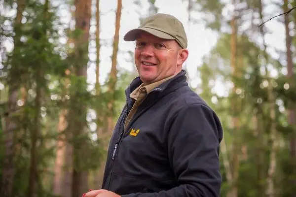 Ein Mann lächelt in einem Wald, trägt eine Mütze und eine Jacke