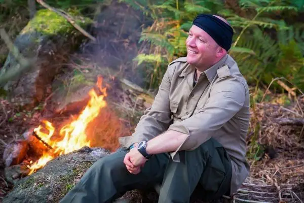Person sitzt lächelnd neben einem Feuer im Wald