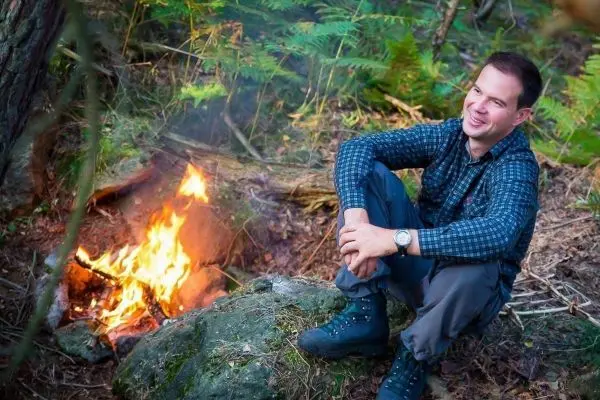 Person sitzt neben einem Feuer auf einem Stein im Wald