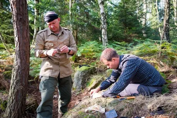 Zwei Personen arbeiten im Wald an einer Feuerstelle mit Werkzeugen und Zunder