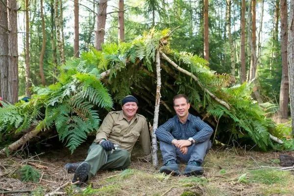 Zwei Personen sitzen entspannt vor einer selbstgebauten Tarp-Notunterkunft aus Ästen und Farnen
