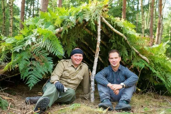 Zwei Personen sitzen vor einer selbstgebauten Tarp-Notunterkunft aus Ästen und Farnen