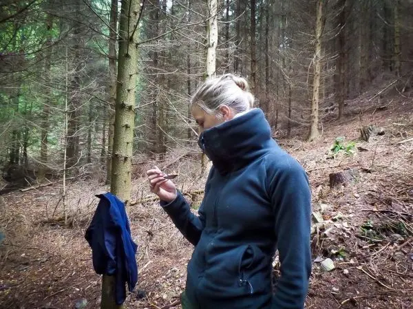 Frau hält einen Gegenstand in der Hand, steht im Wald zwischen Bäumen