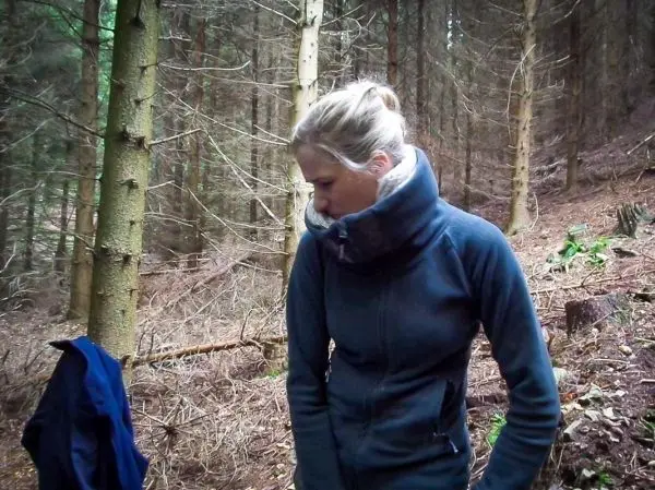 Frau in blauer Jacke steht im Wald zwischen Bäumen und schaut nach unten