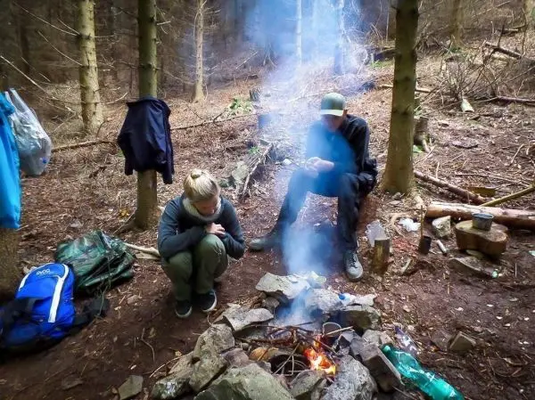 Kleine Flamme in einem Steinkreis, Rauch steigt auf, zwei Personen sitzen daneben