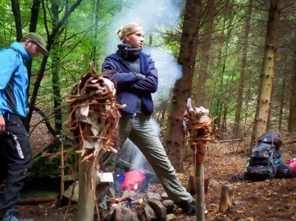 Kleine Flamme raucht zwischen zwei Holzstämmen, im Hintergrund Rucksäcke