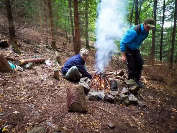 Kleines Bodenfeuer mit Rauch in einem Waldgebiet, zwei Personen in der Nähe