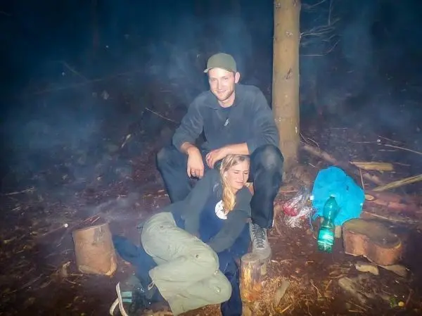 Kleines Bodenfeuer mit Rauch, zwei Personen sitzen auf Holzstämmen