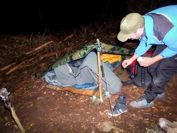 Mann bereitet Tarp-Notunterkunft mit Schlafsack und Stöcken vor