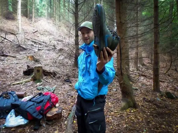 Mann hält einen Schuh in der Hand, im Hintergrund Rucksäcke und Wald